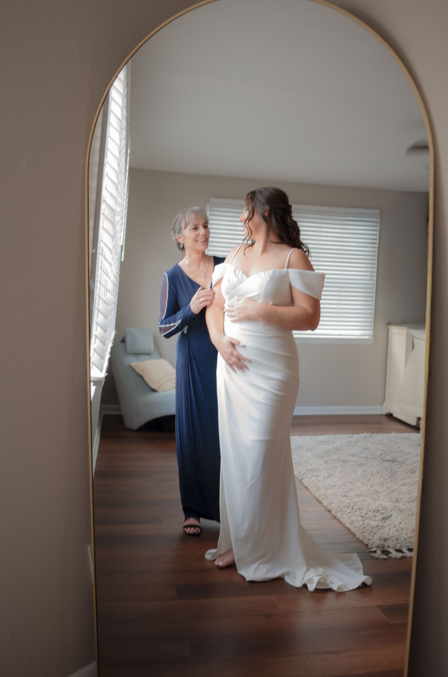 Bride in white off-shoulder gown with older woman in blue dress helping her prepare in a bright room.