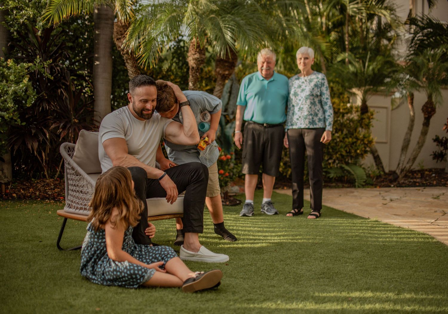 Multi-generational family gathering on a lawn with palm trees and tropical landscaping in the background.