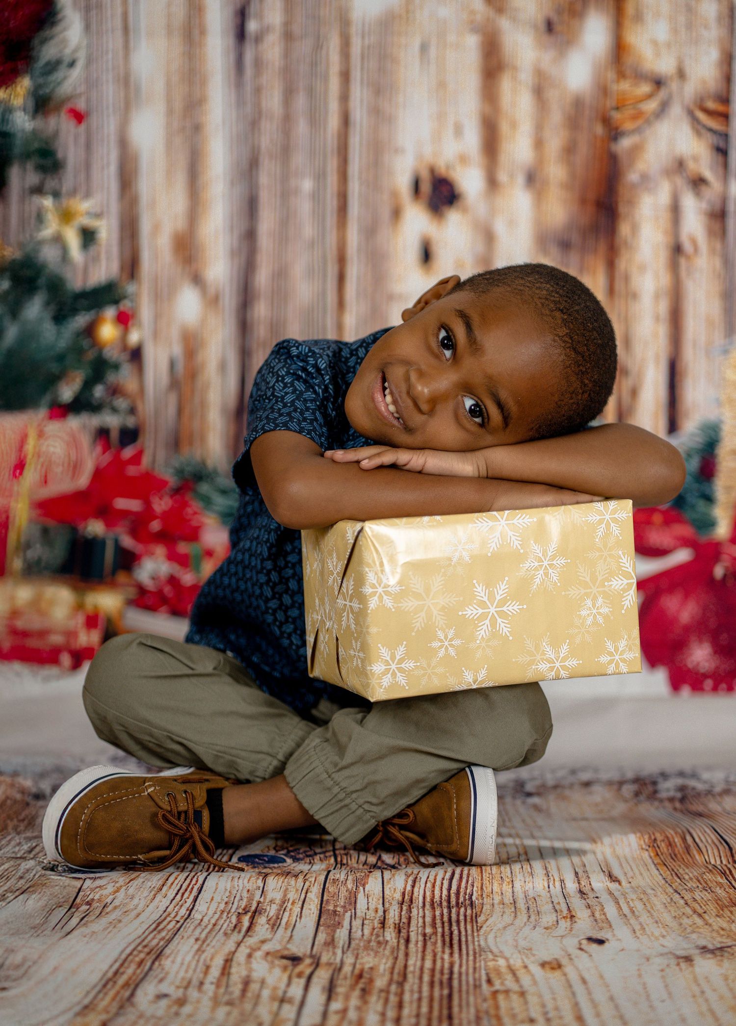 Young child lying playfully on a gold wrapped Christmas present against a rustic wooden backdrop.