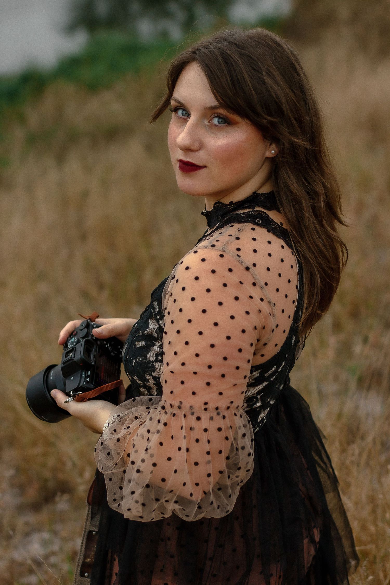 A photographer wearing a sheer polka dot blouse and dark vest poses with a camera in a rustic outdoor setting.