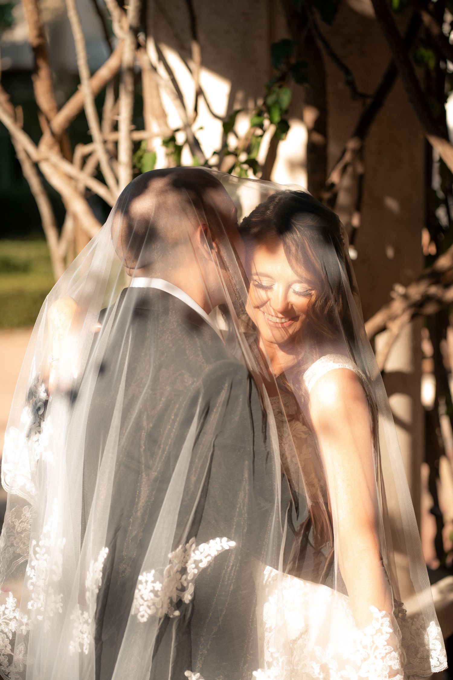 Smiling bride and groom embrace under a sheer lace-trimmed wedding veil in warm golden sunlight surrounded by trees.
