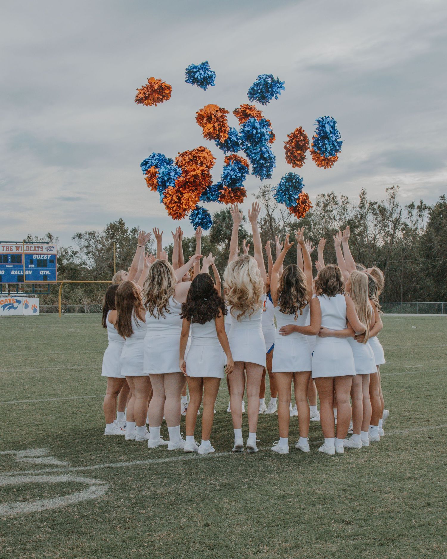 Cheerleading team in white uniforms forming a circle on a sports field with pom poms raised.