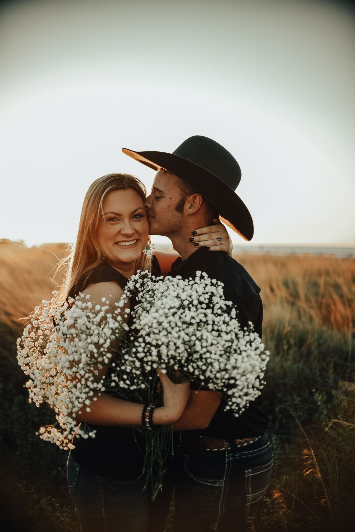 A couple poses with baby's breath flowers in a rustic outdoor setting at sunset.