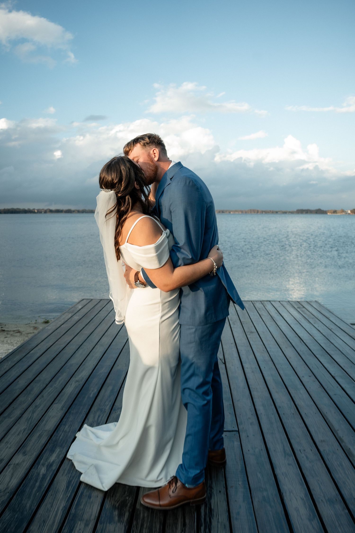 Bride and groom share a romantic kiss on a wooden dock overlooking a serene lake under a blue sky.