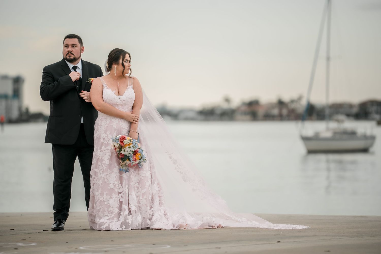 Bride and groom pose on waterfront dock, bride in lace gown holding bouquet, sailboat in background.