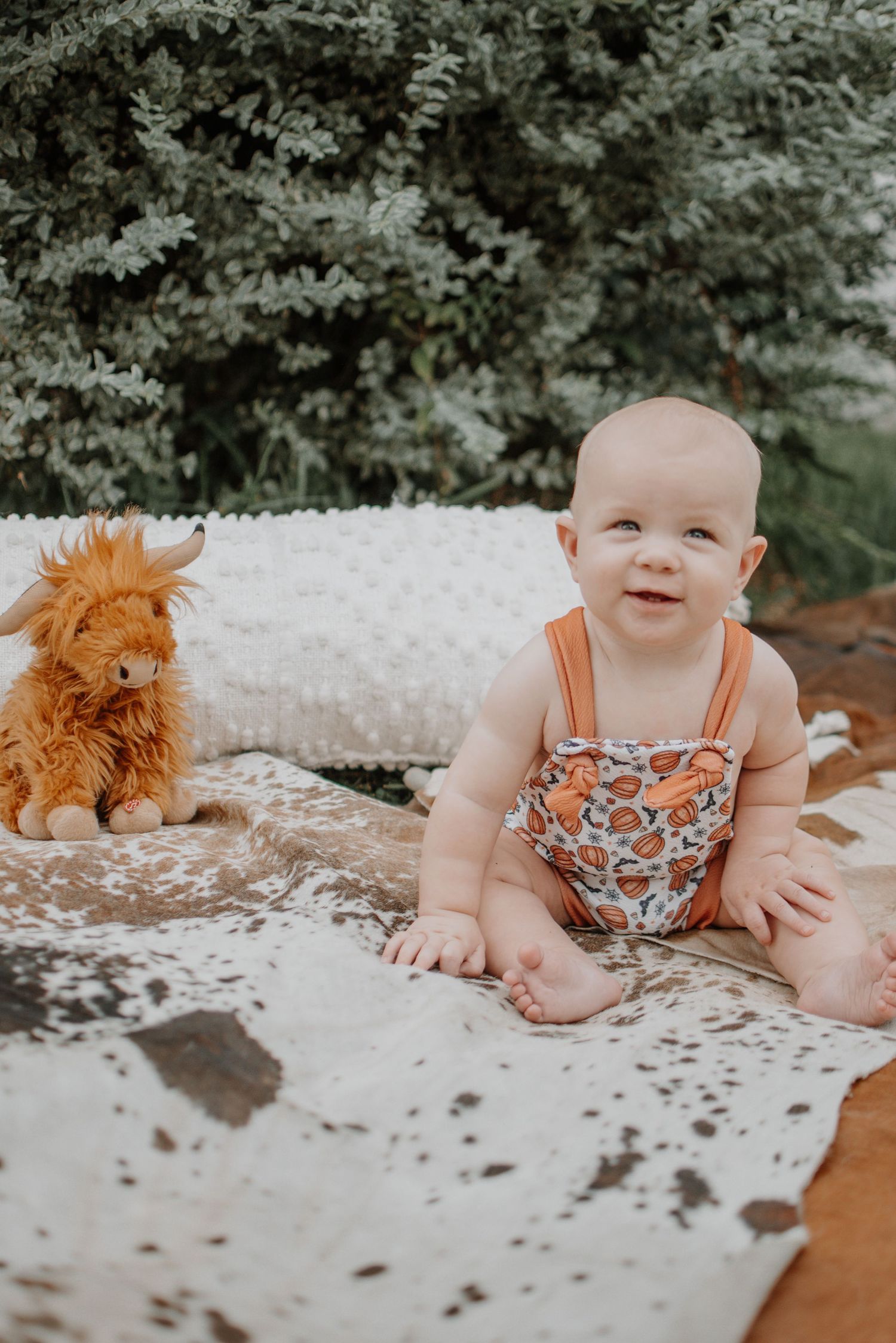Baby in floral romper sitting on blanket with orange plush toy.