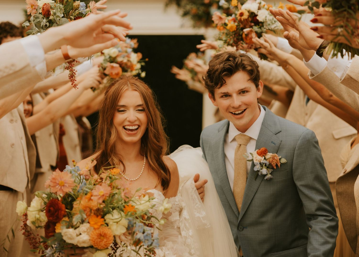 Wedding party celebrates with raised arms in a joyful tunnel exit while holding orange and white floral bouquets.