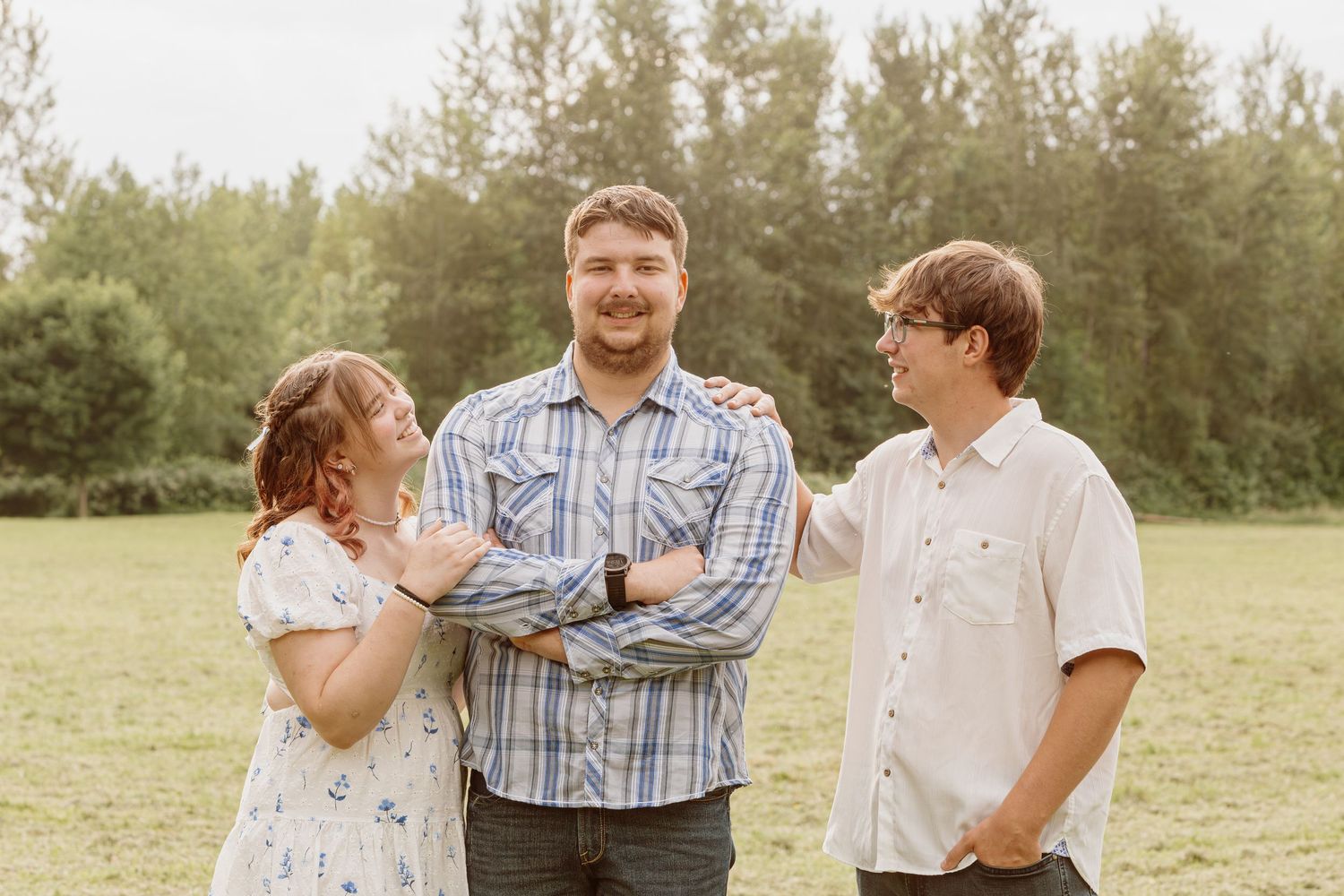 Three siblings laughing and posing together in a green grassy field on a sunny day.