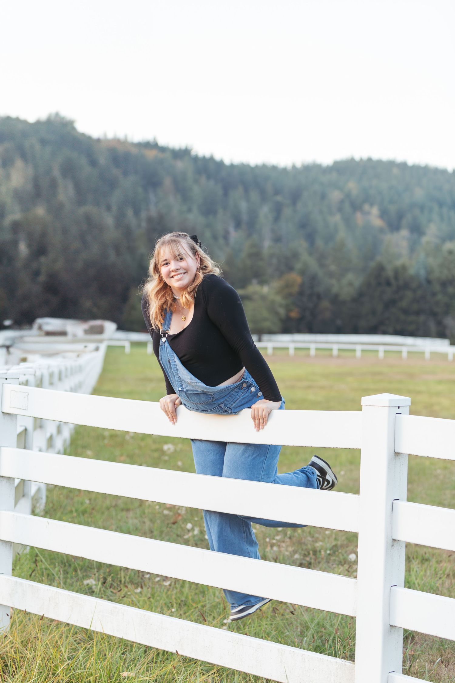 Cheerful young woman in denim overalls leaning on a white farm fence with green hills.