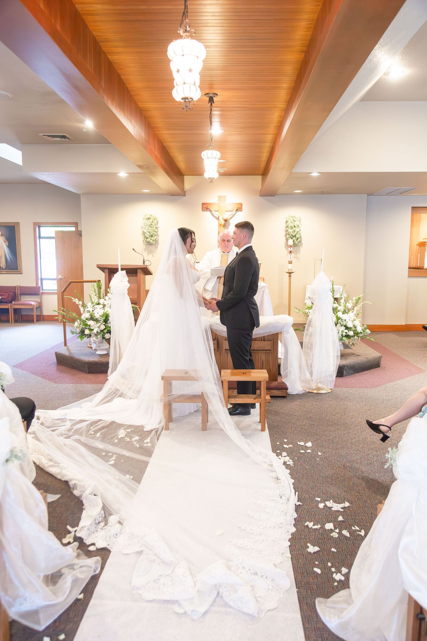 Bride and groom exchange vows at church altar with long white veil trailing down aisle during wedding ceremony.