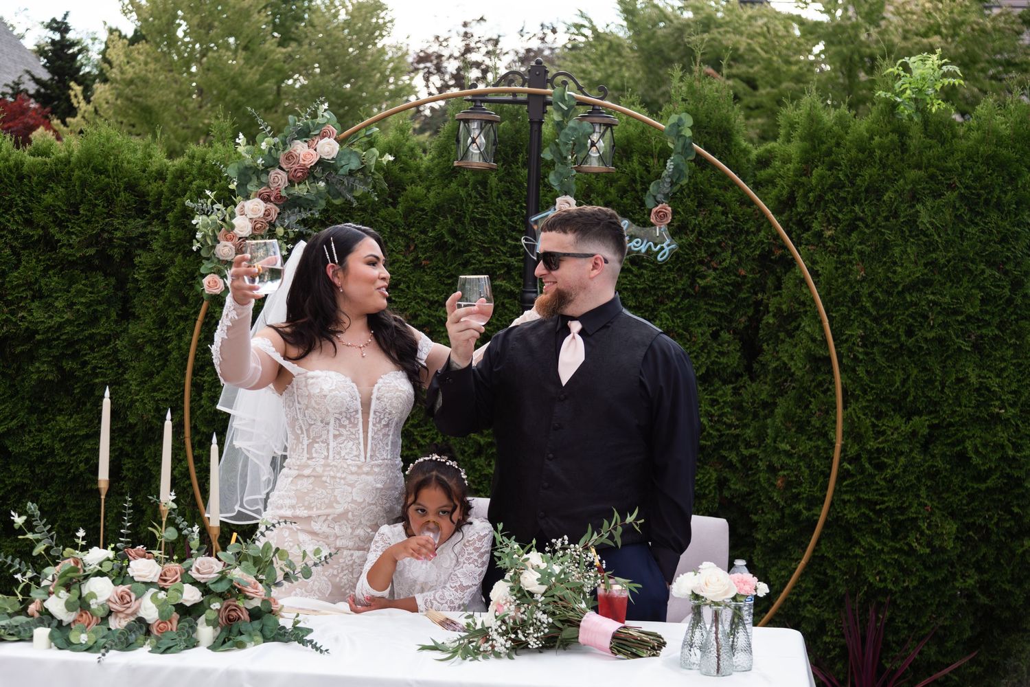 Bride and groom toasting at outdoor wedding ceremony with floral arch, candles, and flower girl at decorated table.