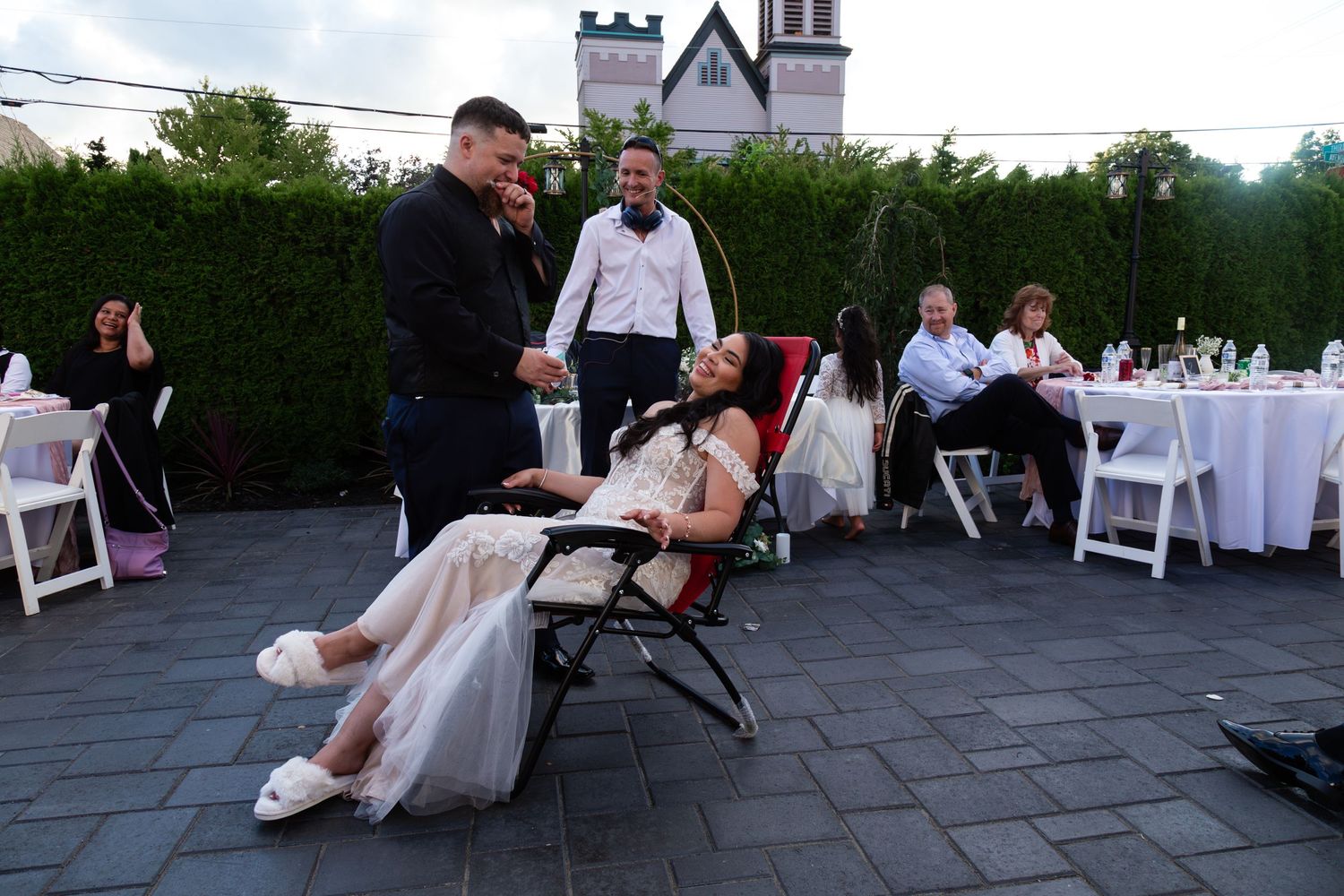 Bride in white gown relaxing in chair surrounded by guests at outdoor evening wedding reception.
