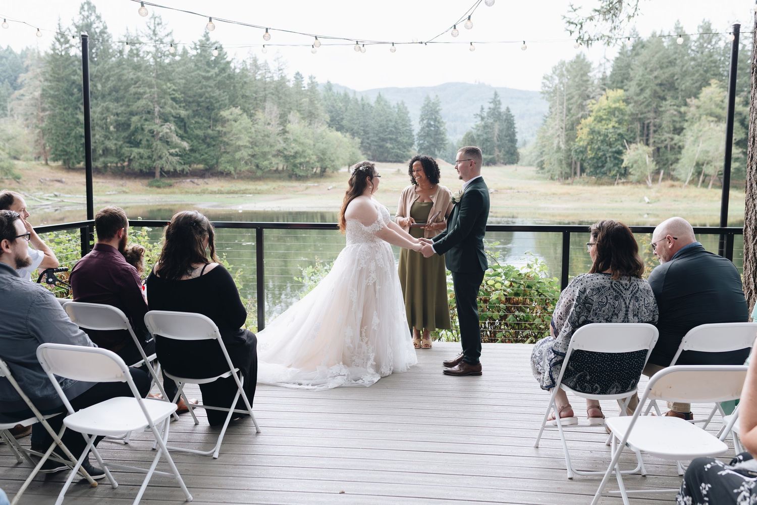 Bride and groom exchange vows on wooden deck overlooking forest pond at intimate wedding.