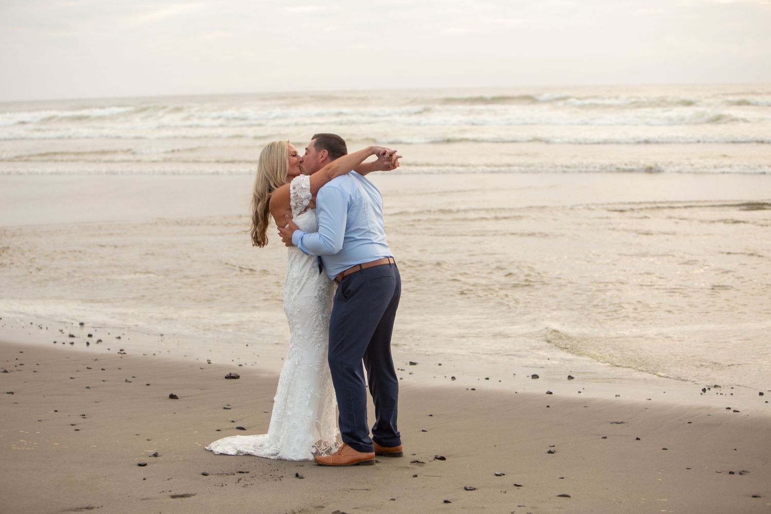 Couple sharing a romantic kiss on a sandy beach in wedding attire, with ocean waves in the background.