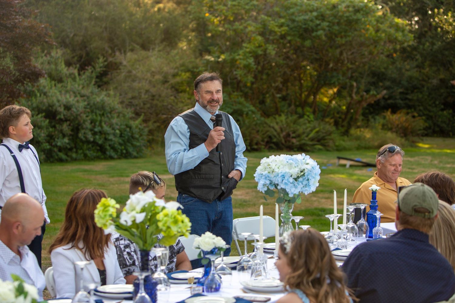 Man giving speech at microphone during elegant outdoor wedding reception dinner with blue floral centerpieces.