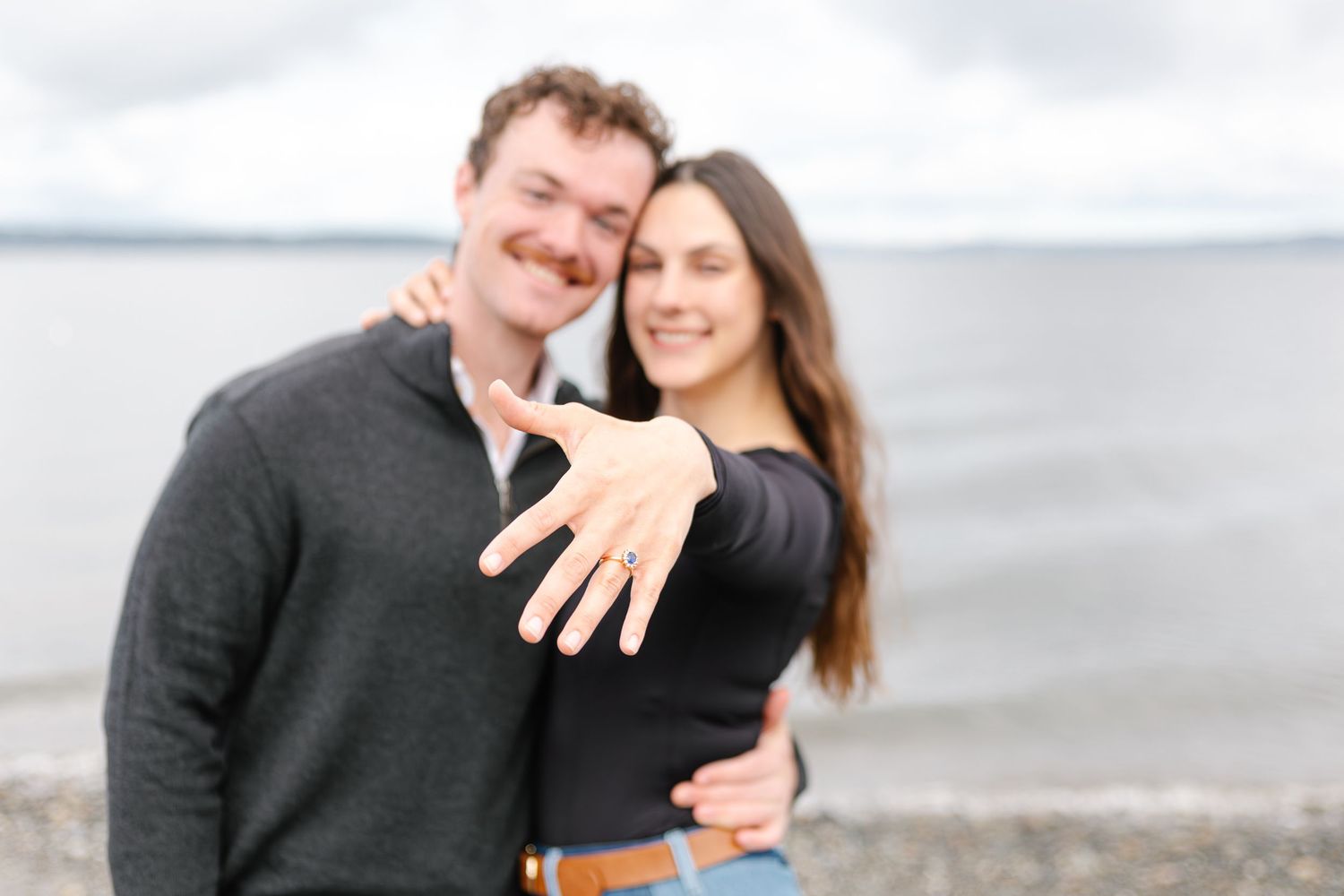 Happy couple showing off engagement ring by the water, woman displaying diamond ring to camera while embracing her fiancé.