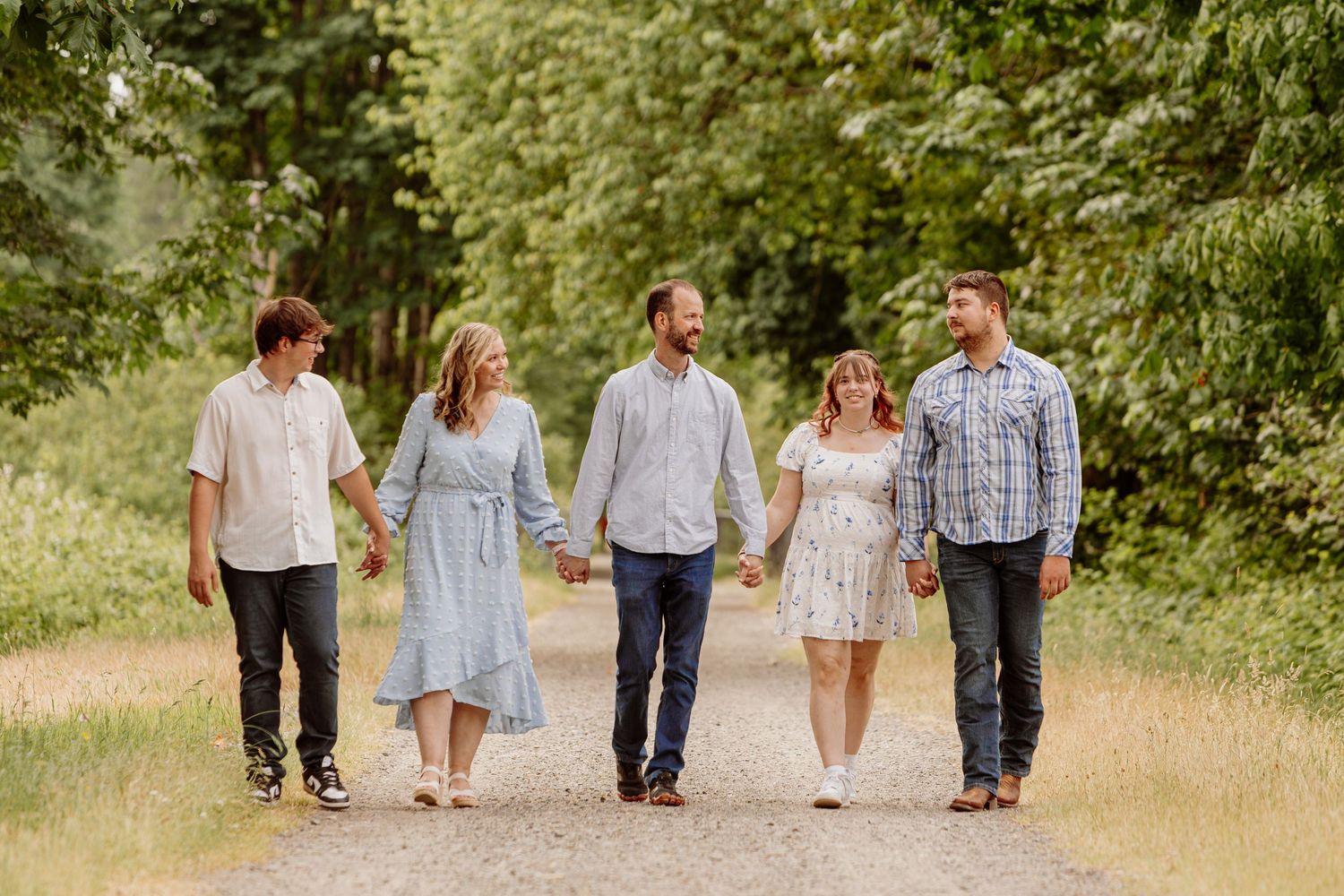 Family of five walking hand-in-hand on a gravel path surrounded by lush green trees on a sunny day.