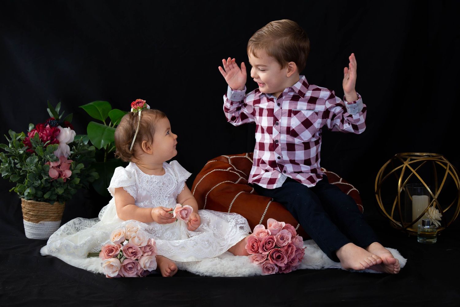 Two young children in formal attire pose playfully against a dark background with pink roses and decorative props.