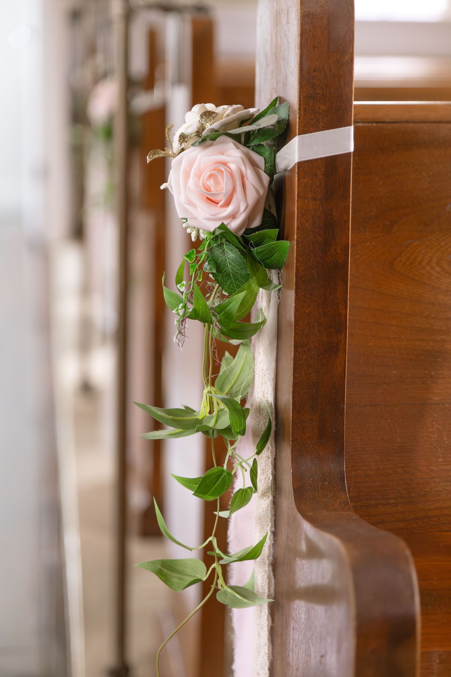 Close-up of a blush pink rose with trailing greenery tied to a wooden church pew with white ribbon.