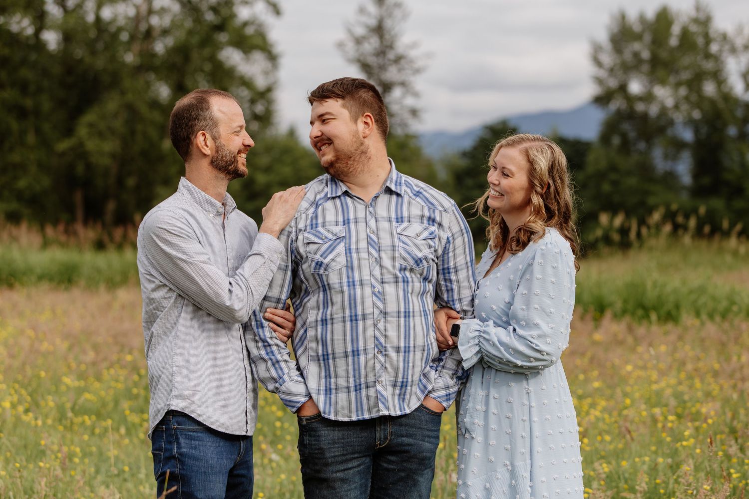 Three people smiling and laughing together in a sunny wildflower meadow with green hills in the background.