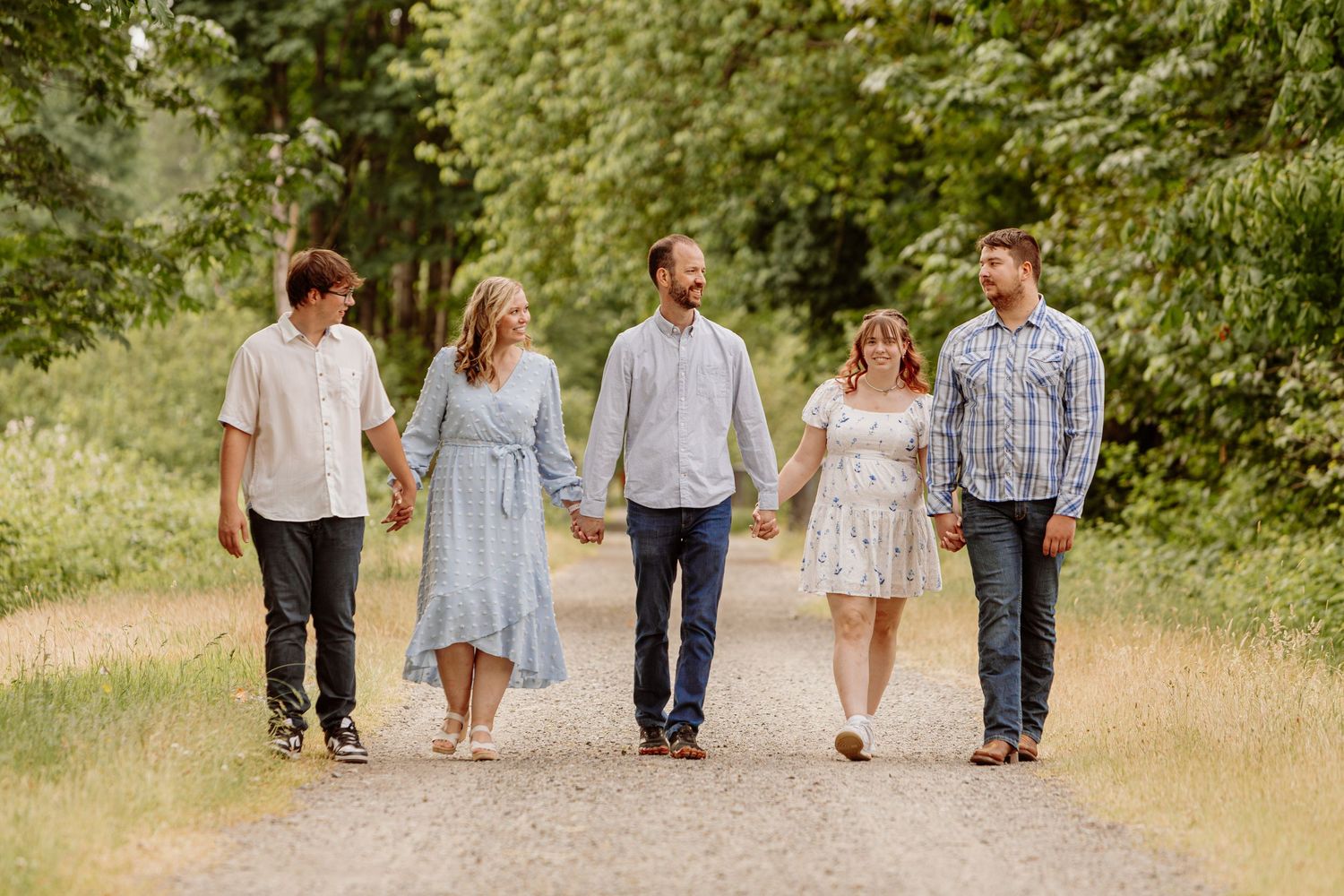 Family of five holding hands walking down a sunlit gravel path surrounded by lush green trees.