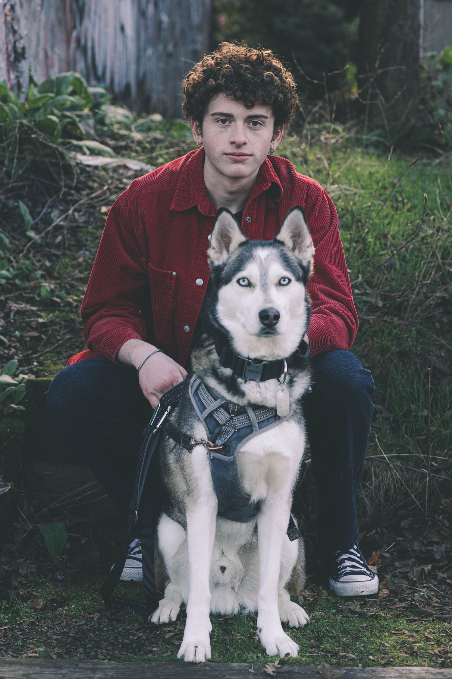Young man in red shirt sitting outdoors with a Siberian Husky dog wearing a harness.