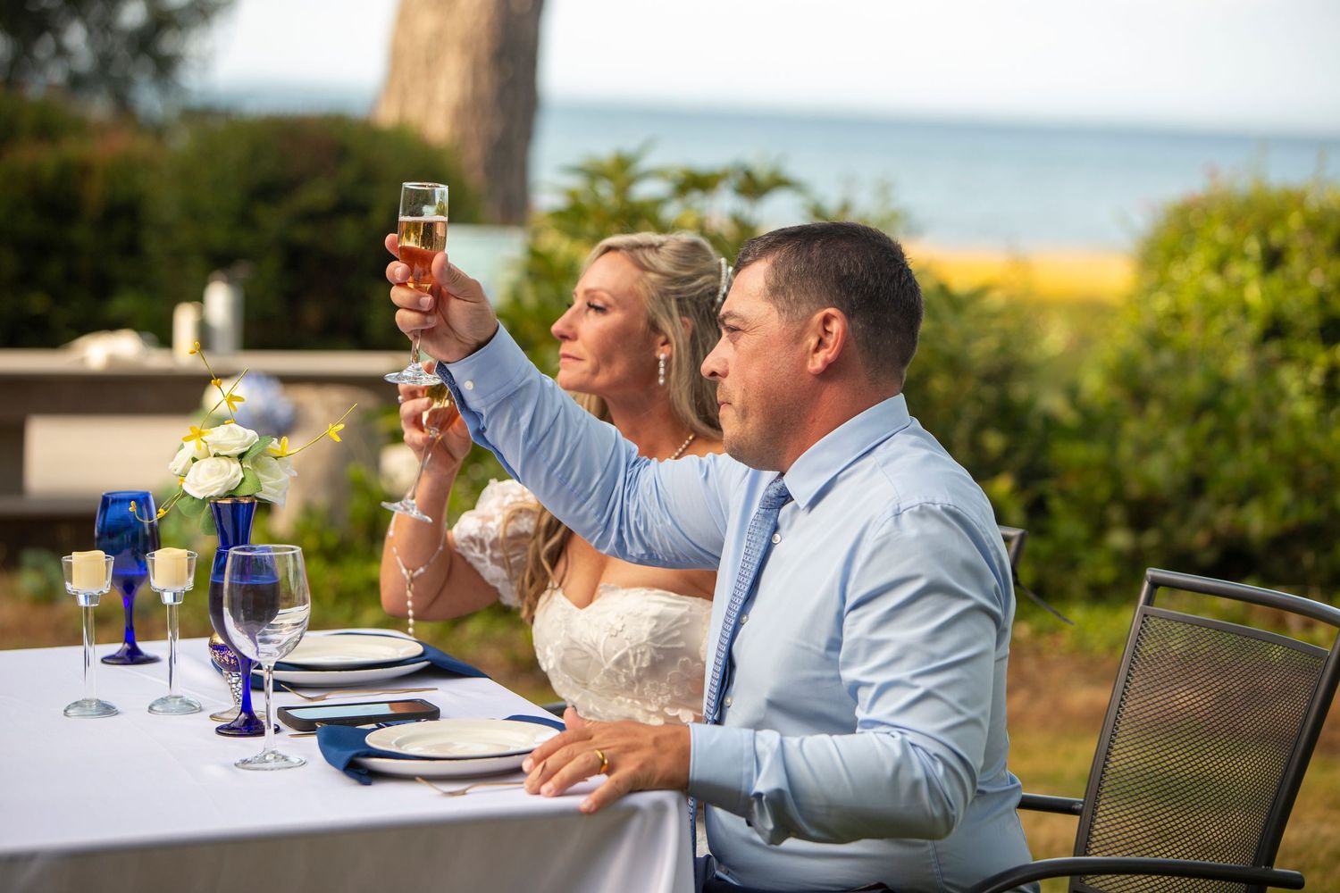 Bride and groom raising champagne toast at outdoor wedding reception table overlooking the ocean.