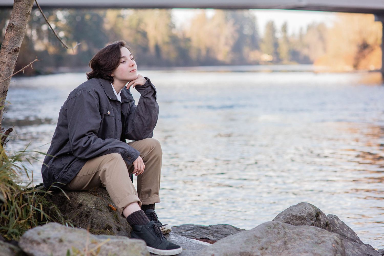 Young woman sitting pensively on rocks by a river with a bridge in the background on a cloudy day.