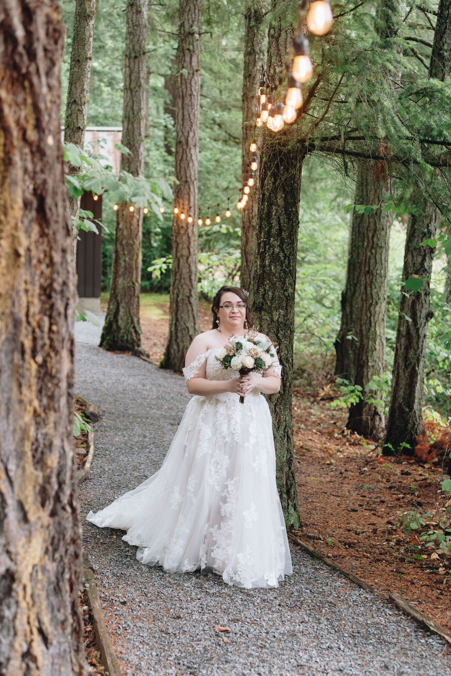 Bride in lace off-shoulder gown holds bouquet on forest path with string lights among tall trees.