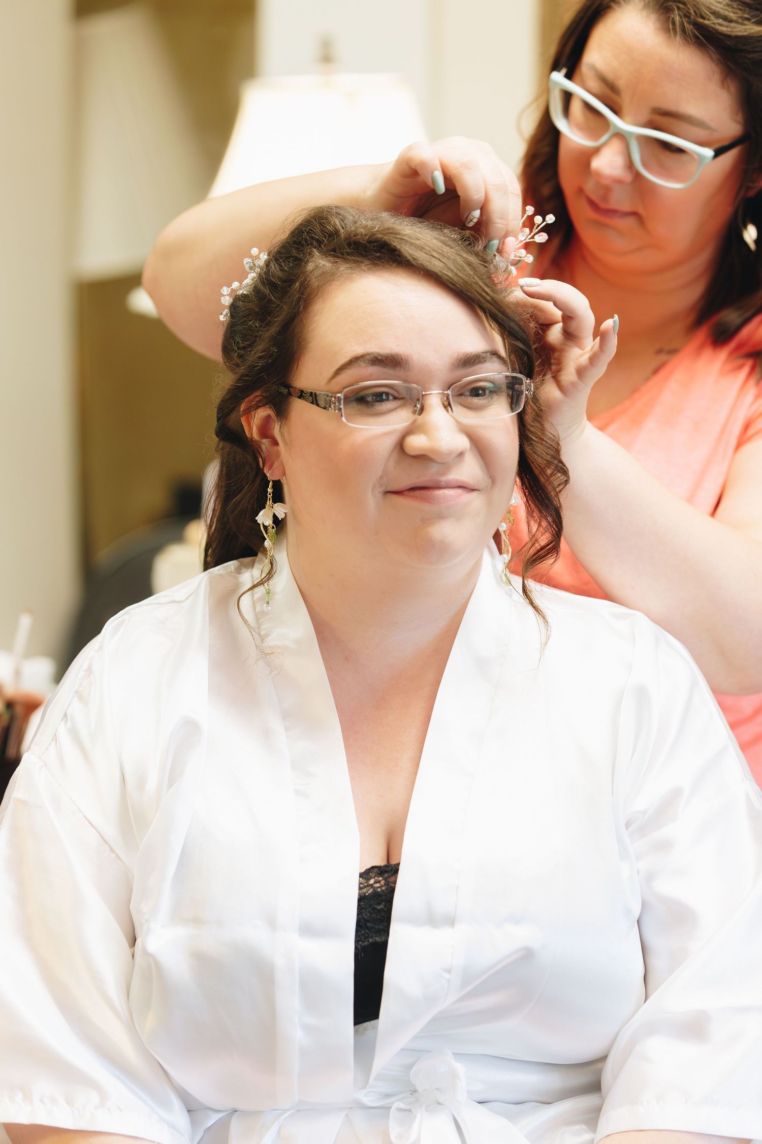Bride in white robe smiling as stylist places floral hair accessory in her hair.
