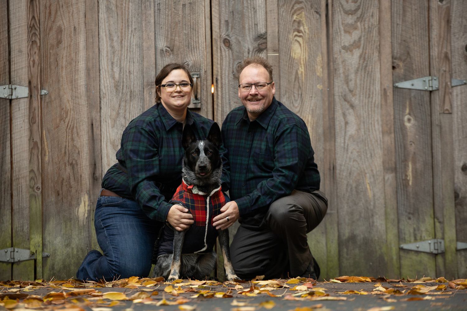 Couple posing with their black and white dog in front of a wooden barn door.