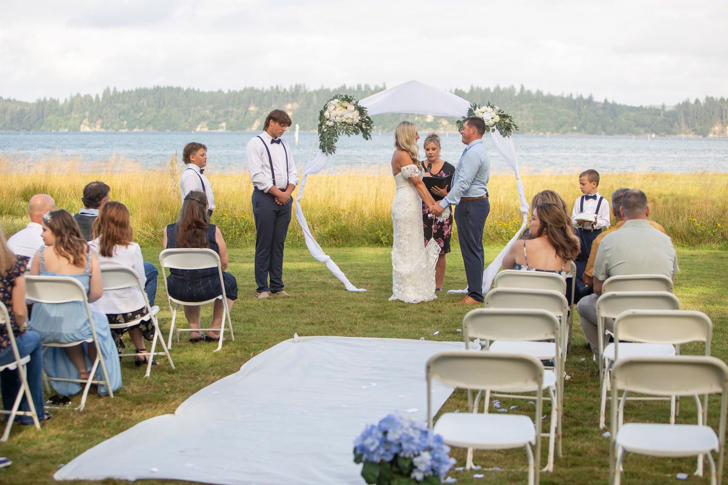 Outdoor waterfront wedding ceremony with white chairs and floral arch on green lawn.