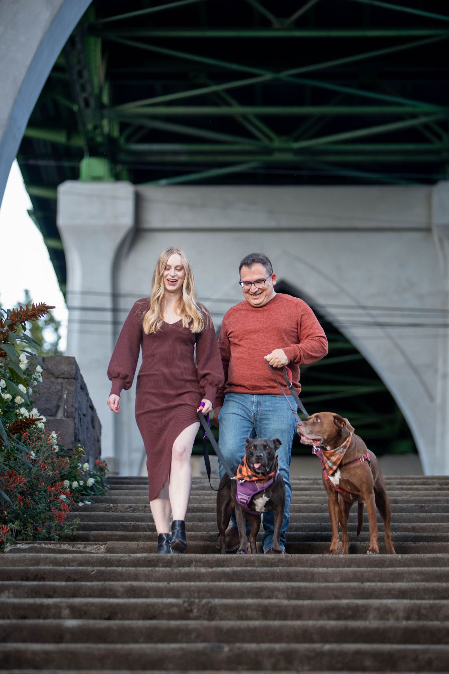 Couple walking two dogs down outdoor steps under a large bridge arch, woman in burgundy dress, man in red sweater.