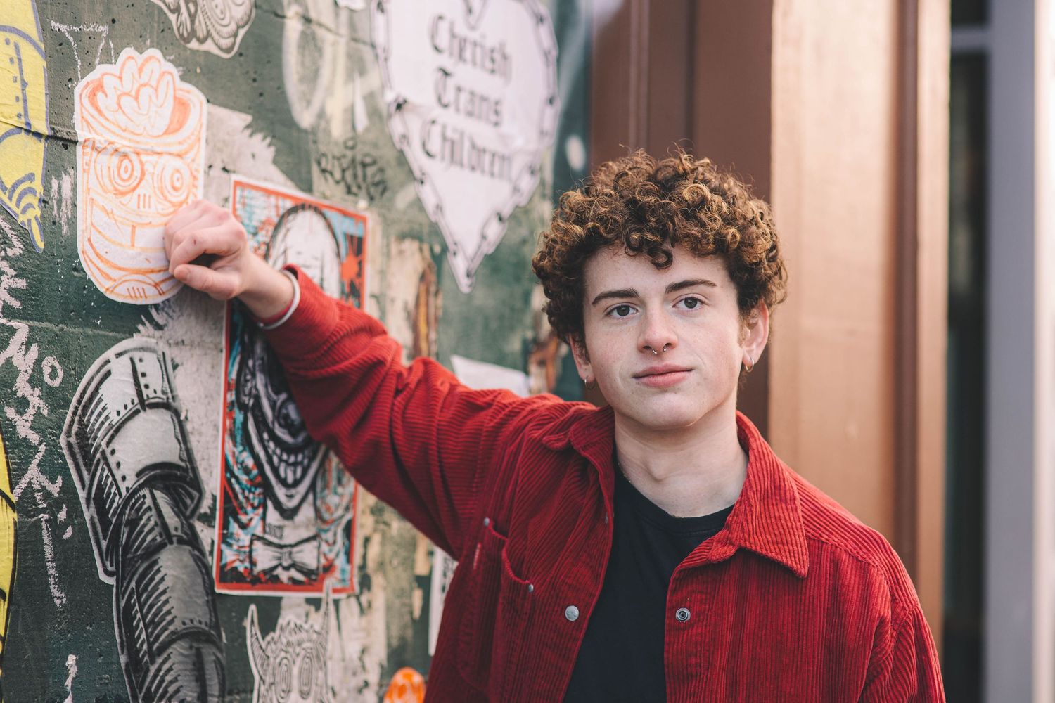 Young man with curly hair in red jacket leaning against colorful street art mural wall.