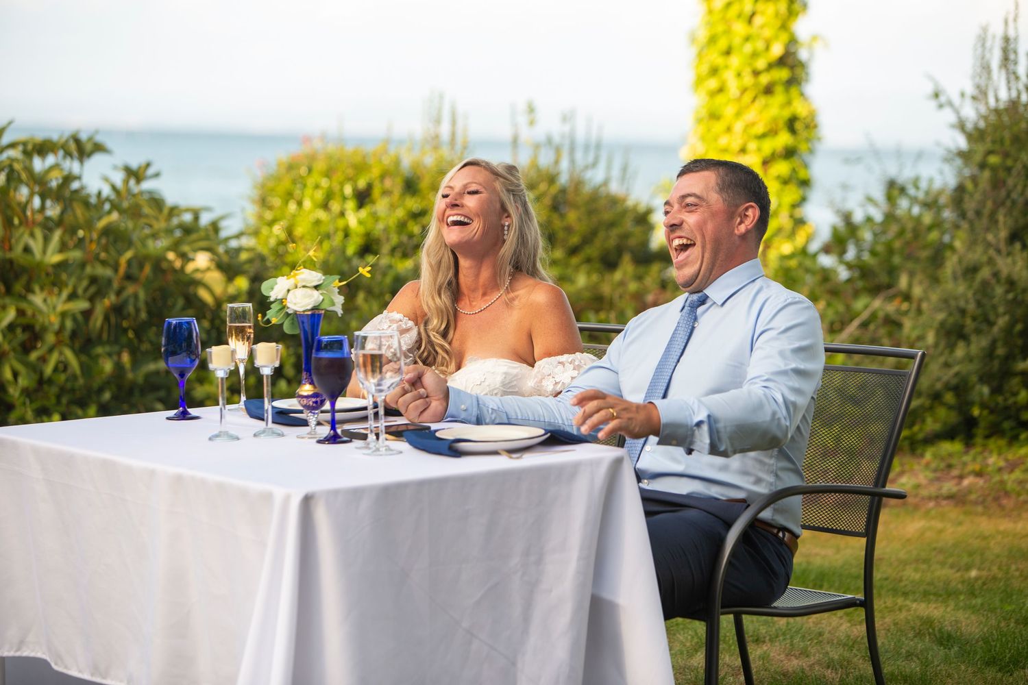 Laughing bride and groom seated at sweetheart table with ocean view during outdoor wedding reception.