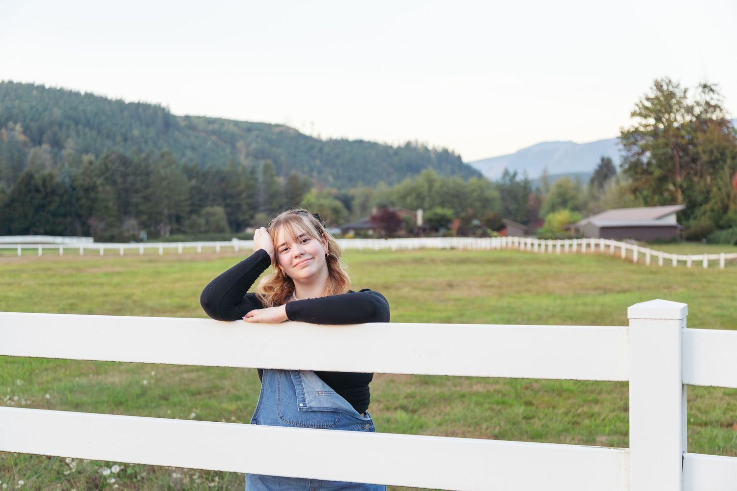 Young woman leaning on white fence at rural farm with green mountains in background.