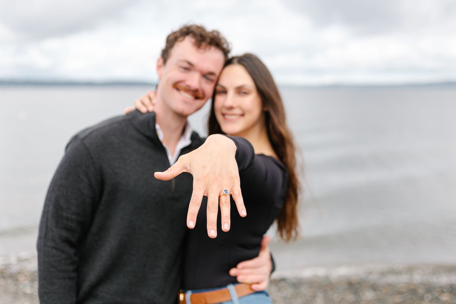 Couple smiling outdoors near water, showing off engagement ring on outstretched hand in a joyful proposal announcement photo.