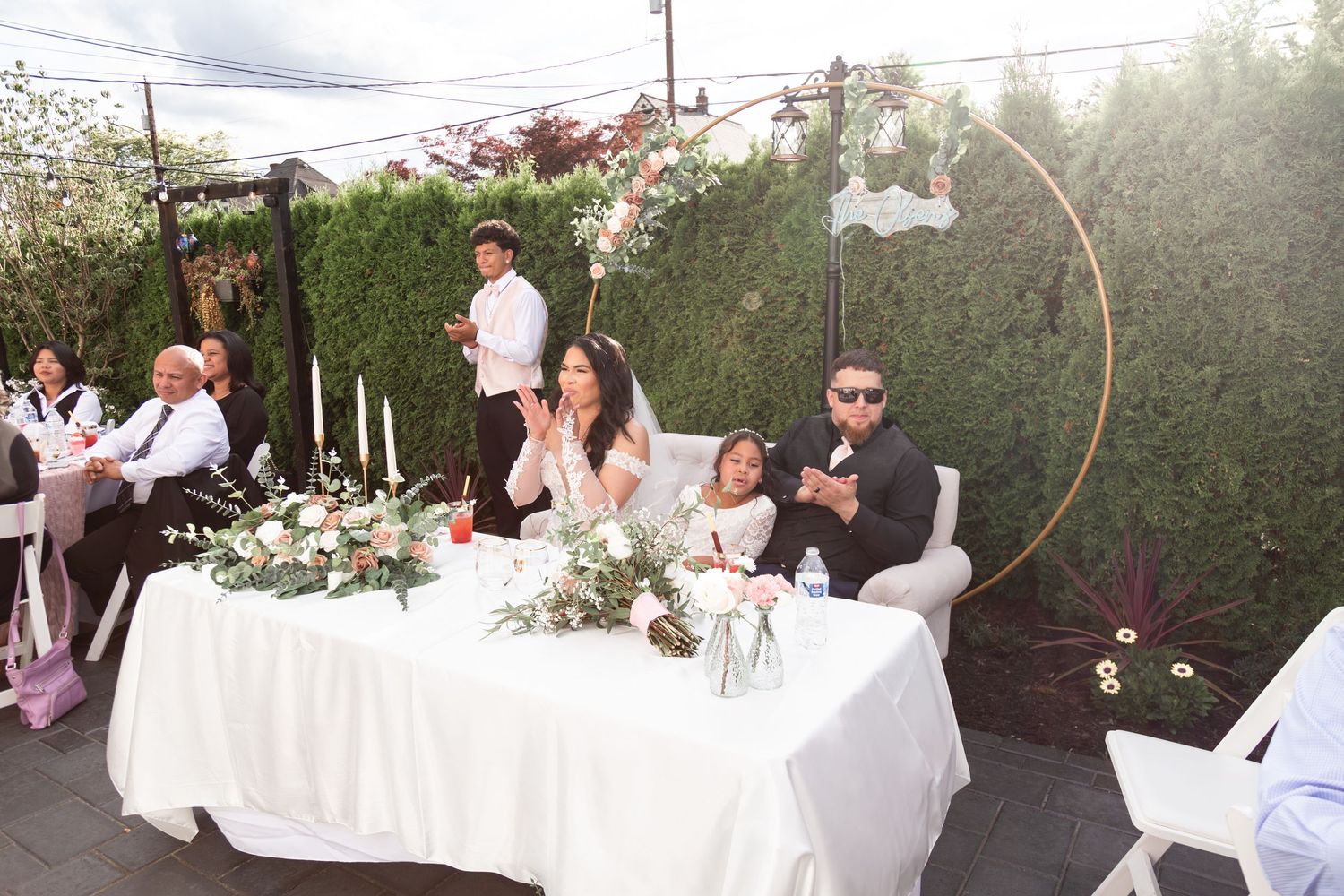 Bride and groom seated at sweetheart table during outdoor wedding reception with floral arch backdrop.