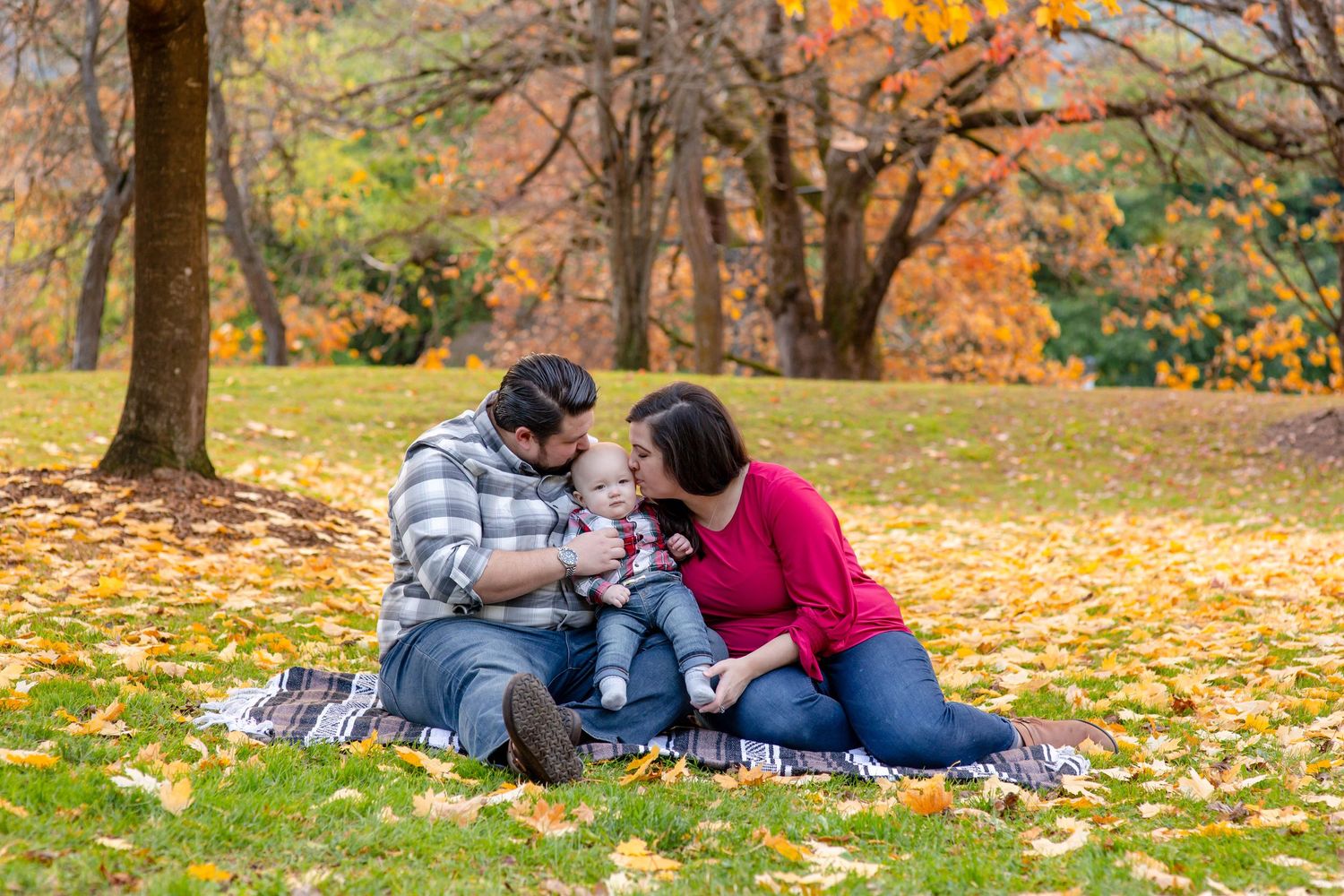 Family sitting on blanket in autumn park, parents kissing baby surrounded by colorful fall foliage.