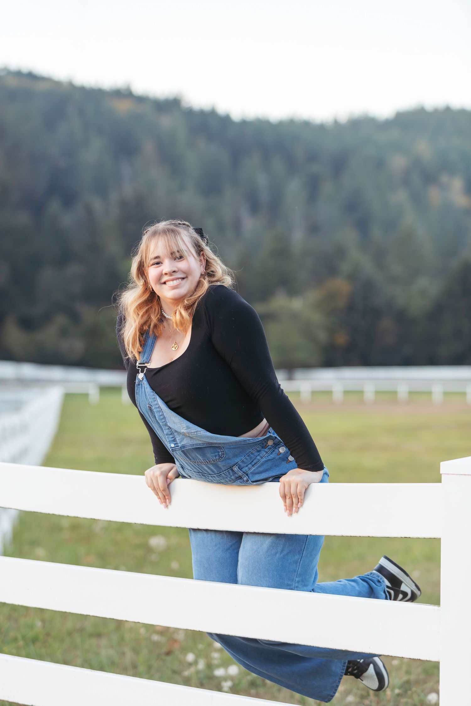 Smiling young woman in denim overalls leaning on white fence with green forested hills in background.