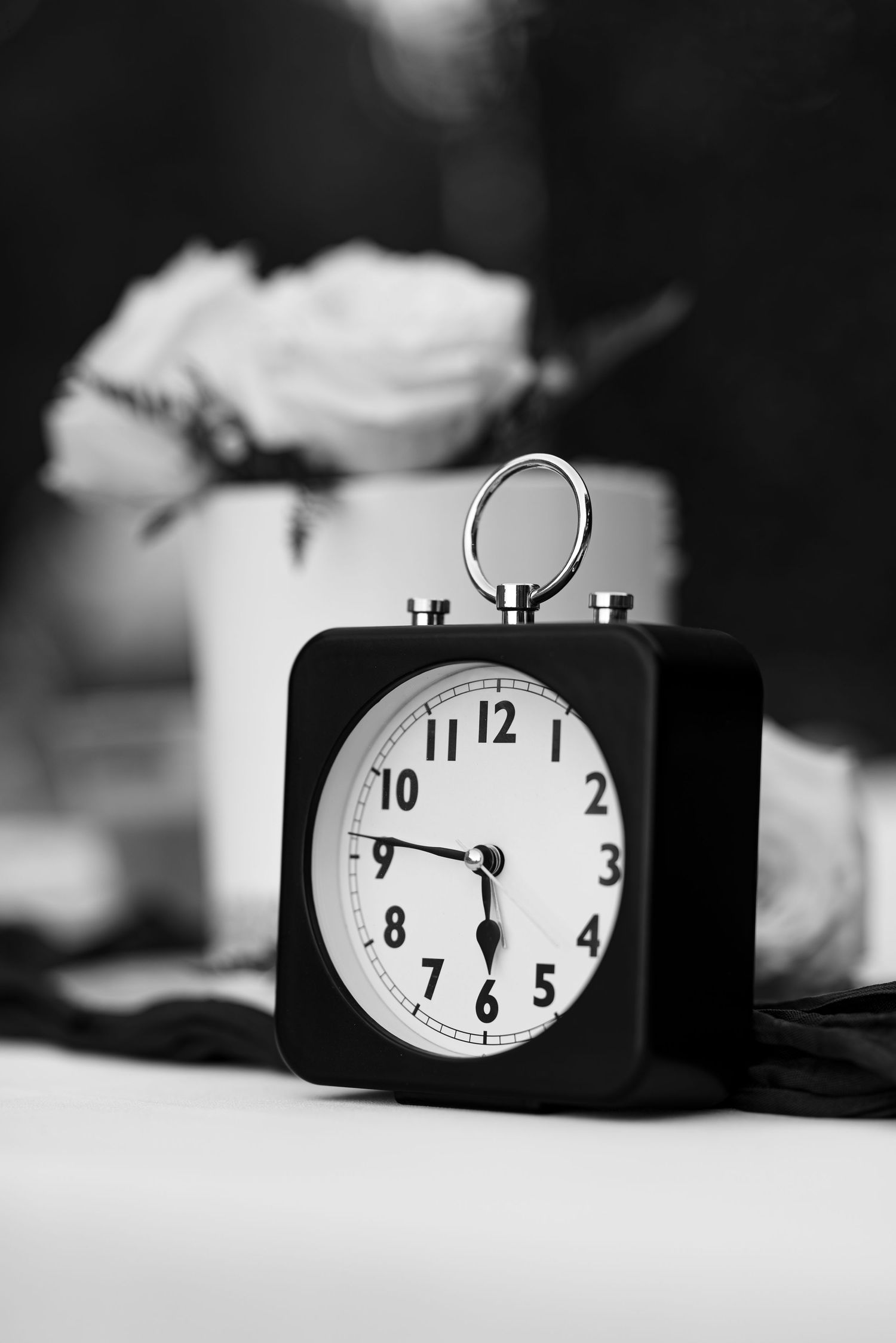 Black and white photo of a vintage square alarm clock showing 6:30 with a white rose in the background.