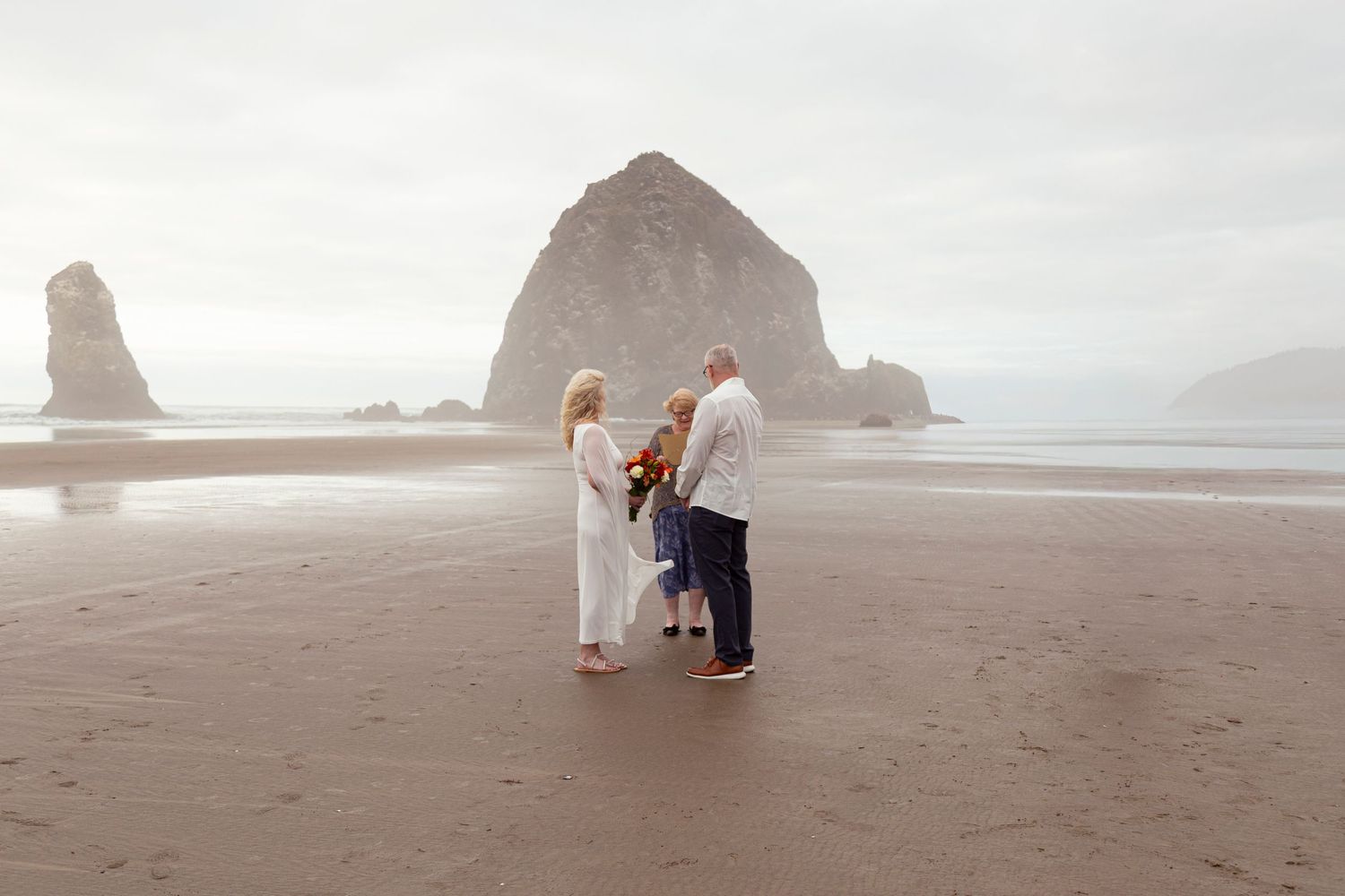 Couple and officiant standing before iconic Haystack Rock during intimate beach wedding.