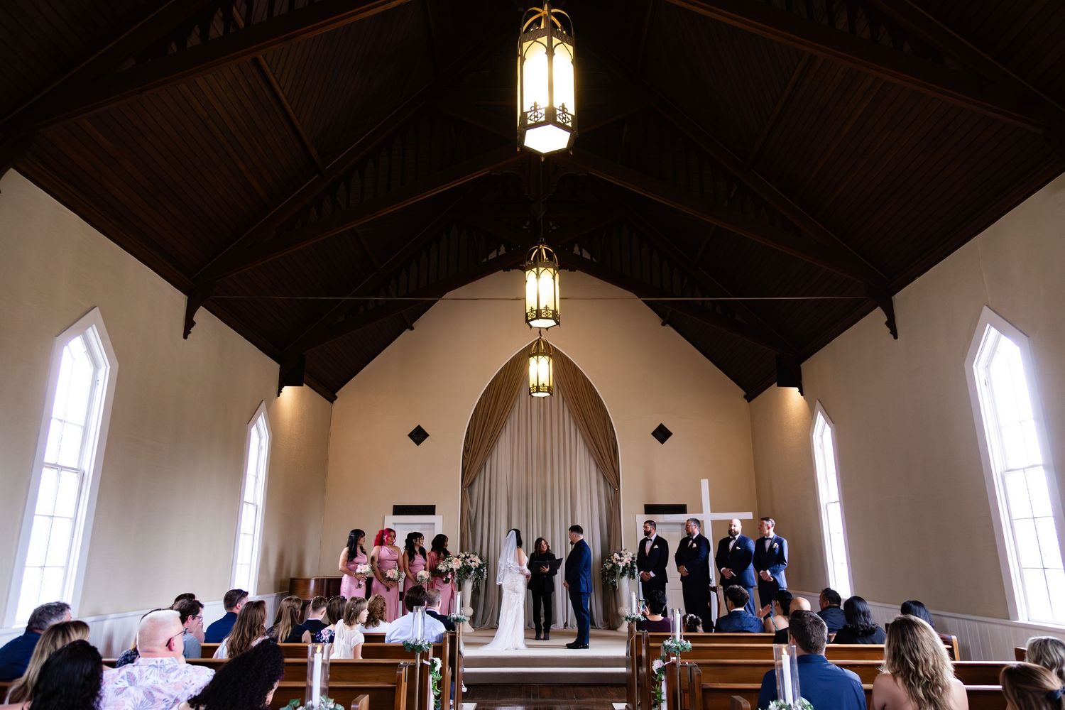 Wedding ceremony in a charming white chapel with wooden pews, arched windows, and elegant hanging lanterns.