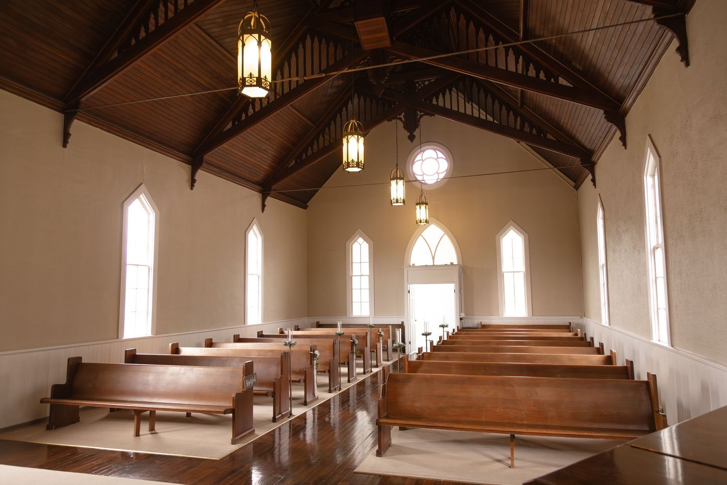 Interior of a charming small chapel with wooden pews, vaulted ceiling, hanging lanterns, and arched windows.