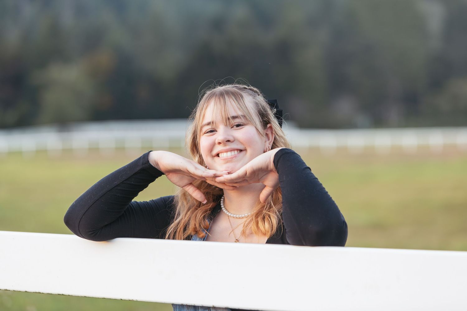 Smiling young woman posing with hands framing her face by a white fence in a sunny outdoor setting.