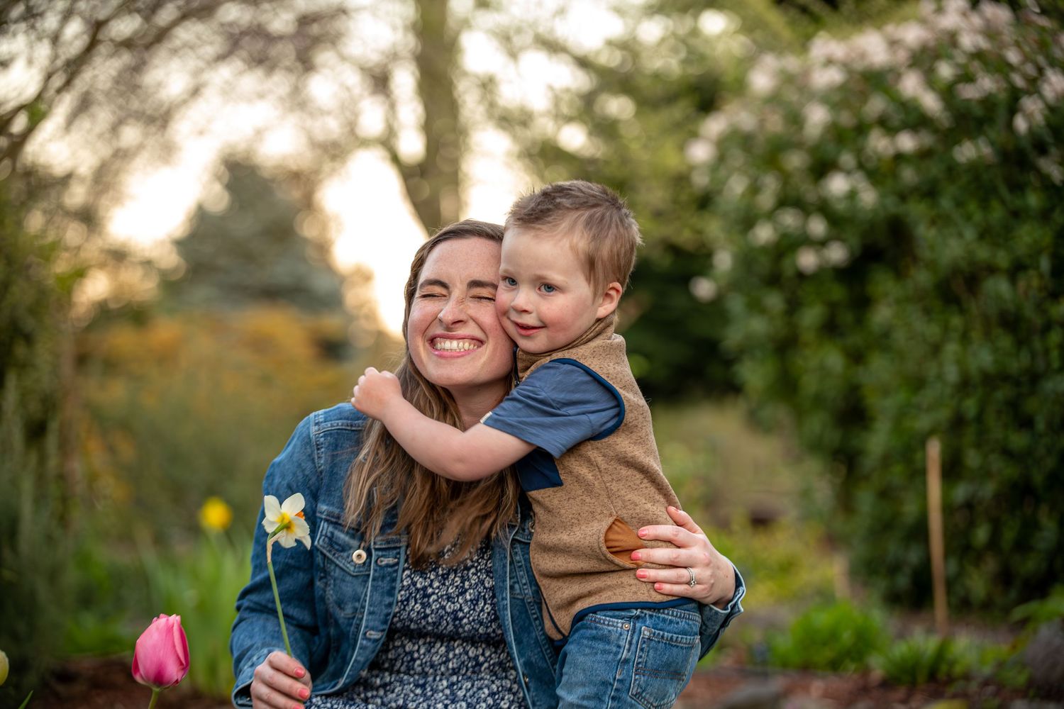 Smiling mother holding young boy who hugs her neck in a lush garden at golden hour.
