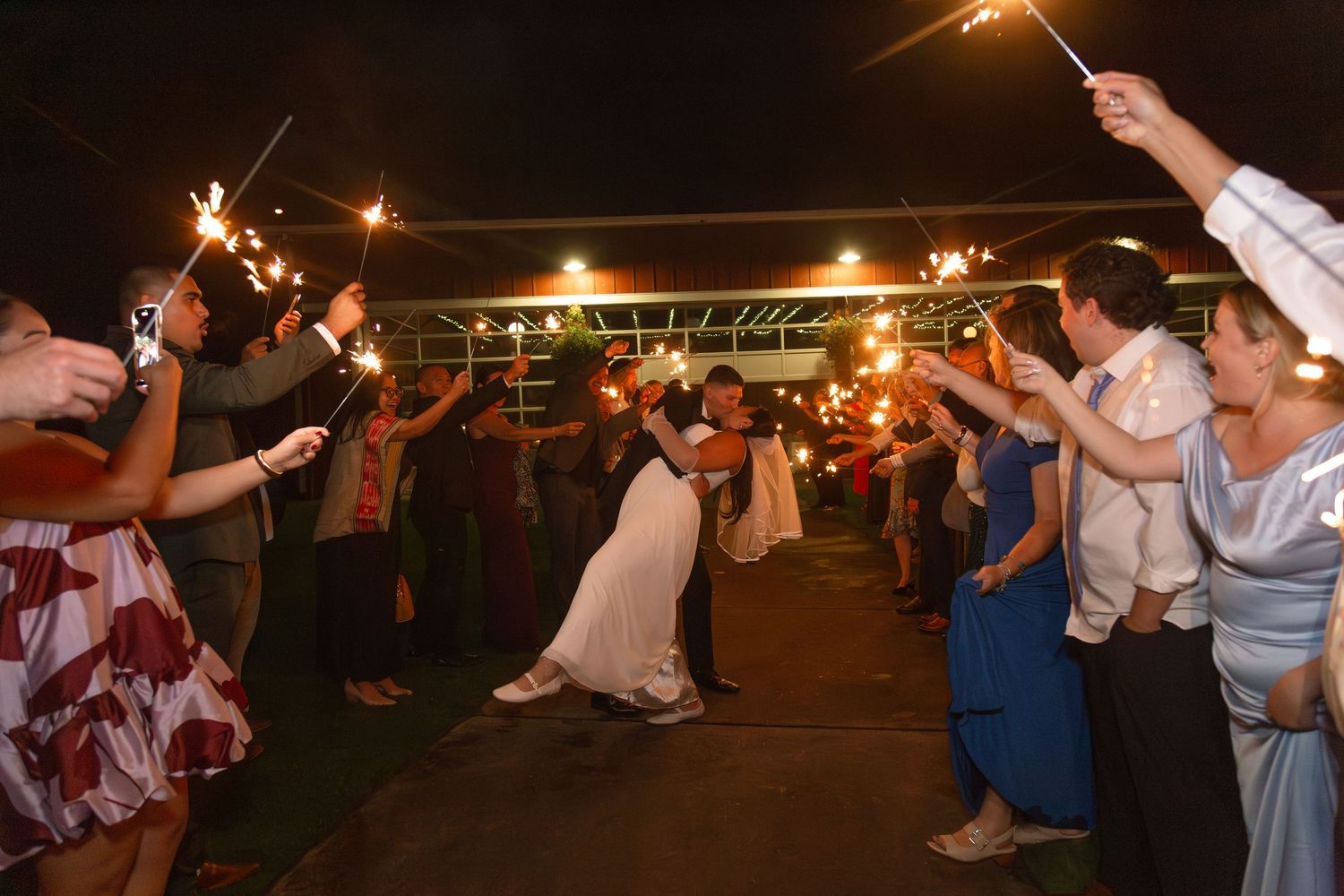 Wedding couple kissing under sparklers held by guests during a romantic nighttime sparkler send-off celebration.