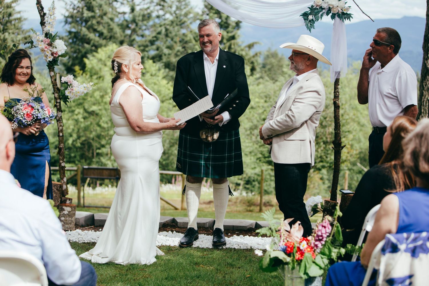 Outdoor wedding ceremony with bride, groom in kilt, and officiant under a floral arch.