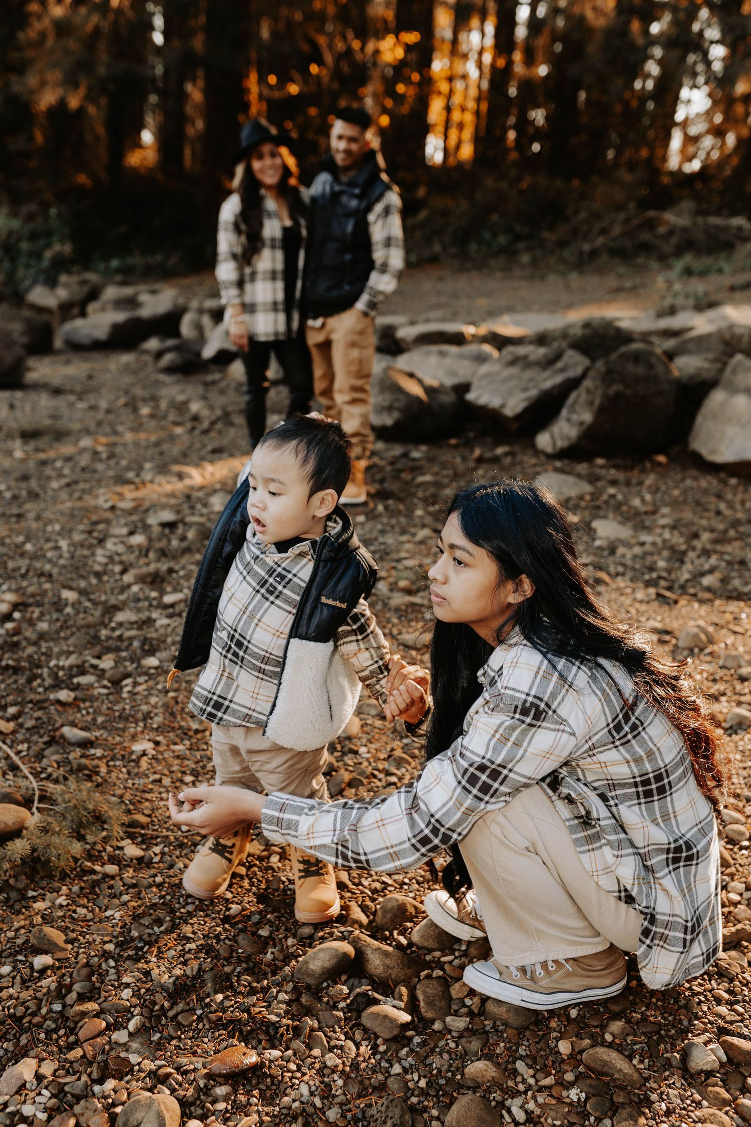 Family in matching plaid shirts exploring a rocky forest trail, with a woman crouching to help a toddler pick up stones.