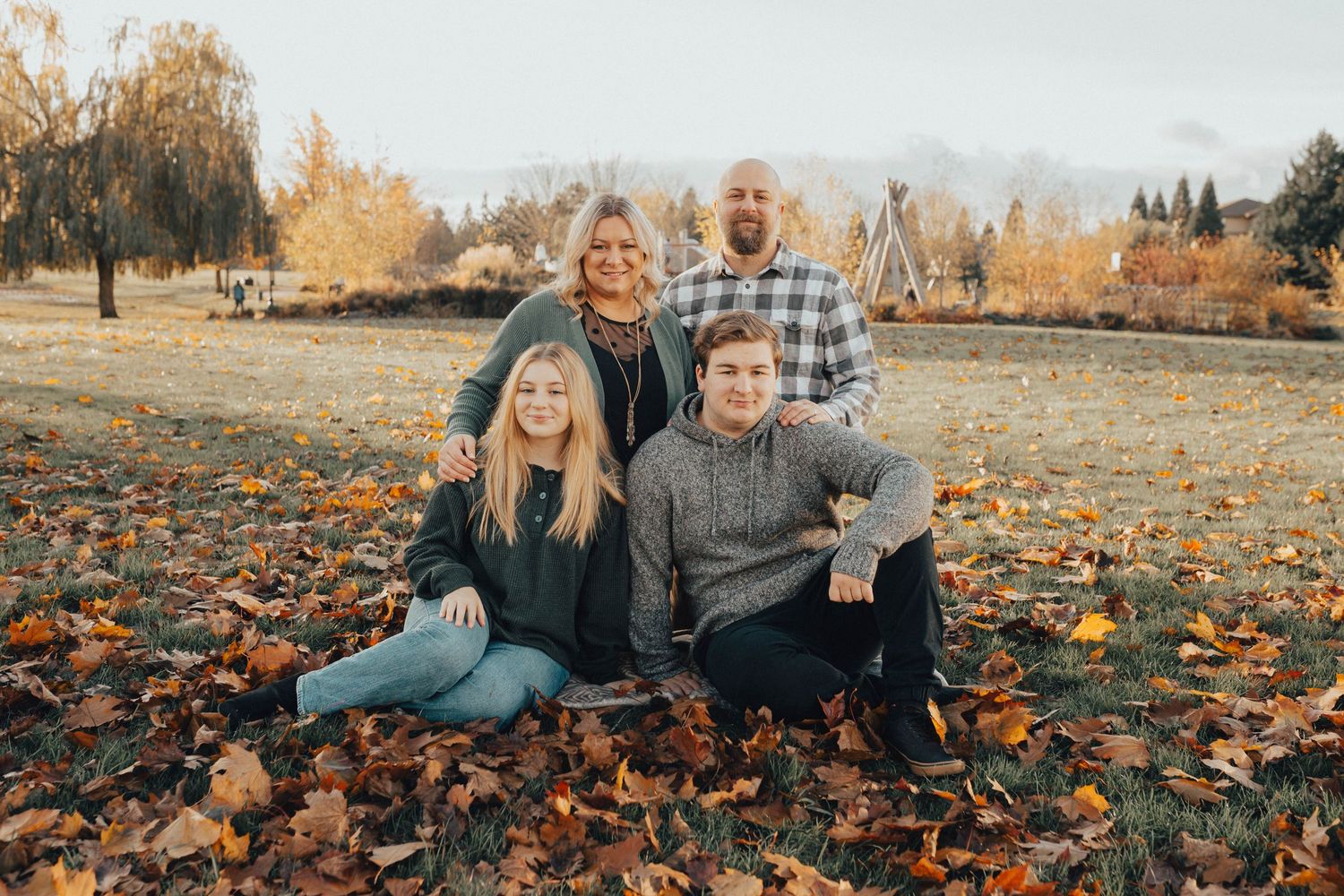 Family of four posing in a leaf-covered field during fall, surrounded by golden autumn foliage.